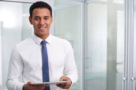 Professional man wearing a white shirt and blue tie standing in a modern glass office holding a tablet, appearing focused and ready to assist
