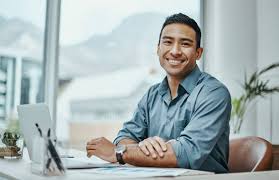 Professional man sitting at a desk in a bright modern office with arms folded, wearing a blue shirt, appearing calm and approachable, with large windows and plants in the background