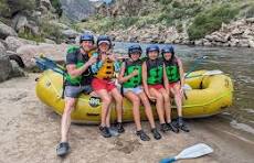 Five people wearing helmets and green life jackets sitting together on a yellow inflatable raft at the edge of a river; the group is smiling and appears happy and excited, surrounded by rocky terrain and greenery under a bright sky; a blue paddle rests on the ground nearby