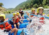 Smiling client wearing a yellow helmet and orange life jacket paddling in a blue raft on a river; water splashes around the group, surrounded by green trees and hills under a bright sky; the atmosphere is energetic and joyful