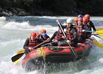 Group of people wearing helmets and life jackets paddling together in a red raft on a fast-moving river; water splashes around them and the group appears excited and joyful; the background features lush green trees and rocky riverbanks under a bright sky