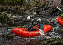 Person wearing a helmet and life jacket paddling alone in a bright orange inflatable raft on a fast-moving river surrounded by rocks and greenery; the individual appears focused and determined as they navigate the rapids; the atmosphere is adventurous and energetic