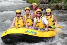 Five people wearing yellow life jackets and helmets smiling together in a yellow raft navigating a fast-moving river surrounded by rocks and greenery; the group appears excited and joyful; the raft has the word ALAN visible on the front