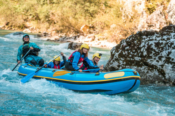Four people wearing yellow helmets and life jackets paddling together in a bright blue inflatable raft as they navigate gentle whitewater on a river. The group appears focused and engaged, surrounded by clear water, rocky banks, and lush green trees in a lively outdoor setting. The mood is adventurous and energetic.
