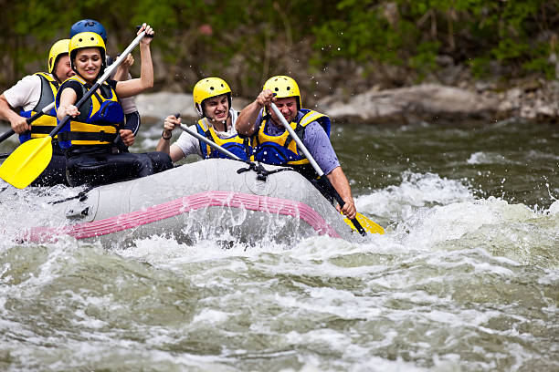 A group of people wearing yellow helmets and life jackets paddling together in a gray inflatable raft as they navigate fast-moving whitewater rapids. The river is turbulent and surrounded by green trees and rocky banks, creating an adventurous and energetic atmosphere.