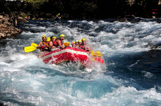 A group of people paddling together in a red inflatable raft as they navigate fast-moving whitewater rapids, all wearing yellow helmets and life jackets. The scene is energetic and adventurous, with churning blue water and rocky riverbanks in a natural outdoor setting. The group appears excited and focused as they tackle the challenging river